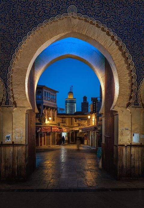 Bab Boujloud Large ornate Moroccan archway framing a view of a narrow street and buildings with a mosque in the background at dusk.