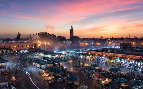 Djemaa al Fna 2 Bustling market square at sunset with stalls, green umbrellas, people, and a mosque tower in the background under a colorful sky.