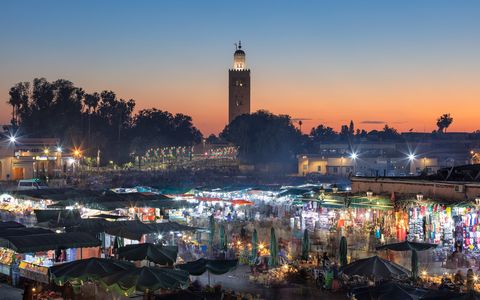 Djemaa al Fna 1 Bustling night market with green canopies, colorful stalls, and bright lights, with a tall minaret and sunset sky in the background.