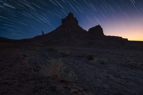 Timeless Rocky desert landscape with scattered shrubs under a night sky filled with star trails and a hint of dawn or dusk on the horizon.